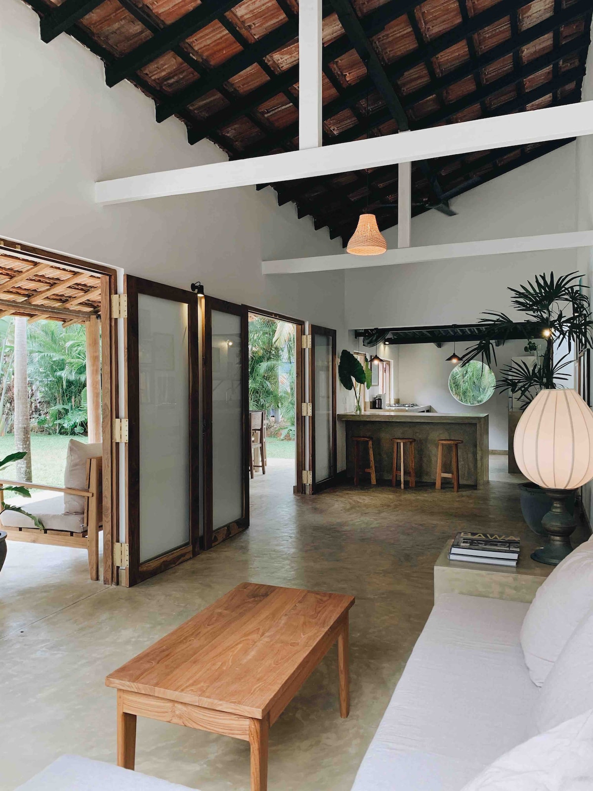 An open living area features a light-colored sofa facing a wooden coffee table. Large glass doors provide a view of the garden, allowing natural light to fill the space. A kitchen area with a bar connects seamlessly to the living room, emphasizing a relaxed flow.