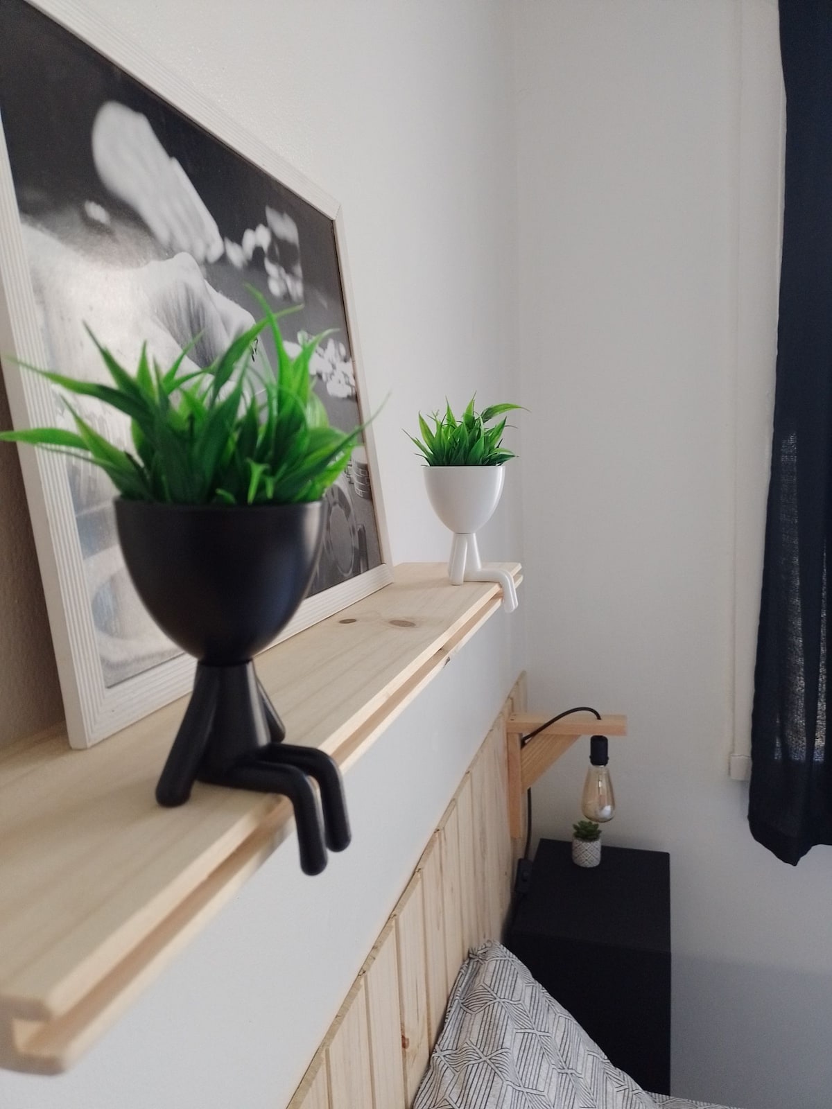 A wooden shelf holds two small plant pots, one black and one white, each adorned with green plants. They are positioned beside a cozy bed, complemented by a nightstand featuring a lamp with an exposed bulb. A framed black-and-white photograph hangs on the wall above.