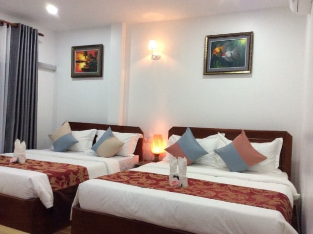 The room features two spacious beds, each adorned with decorative pillows and soft linens. A nightstand with a lamp is situated between the beds, and artwork enhances the bright, neutral walls. Floor-to-ceiling curtains frame windows, adding to the room's inviting ambiance.