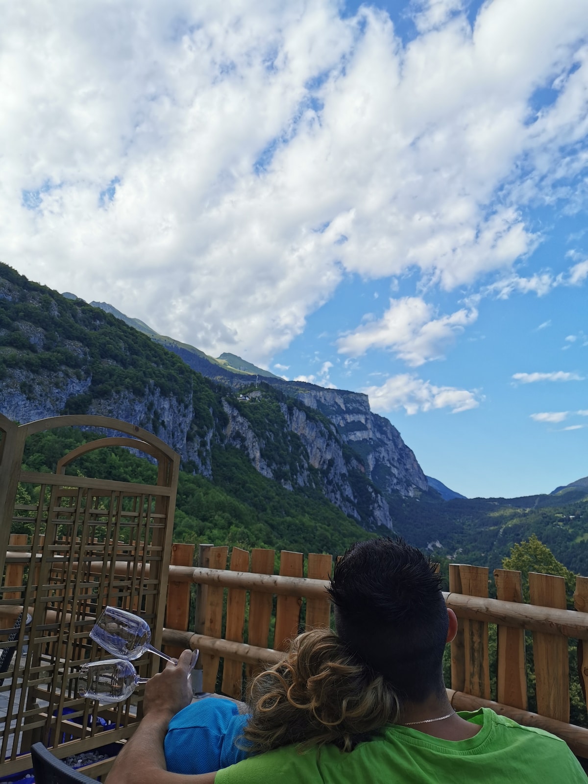 A scenic outdoor terrace is shown, featuring a wooden railing that overlooks lush green mountains and a clear blue sky. The clouds are gently scattered, contributing to the serene ambiance. Comfortable seating is visible, inviting relaxation amidst nature's beauty.