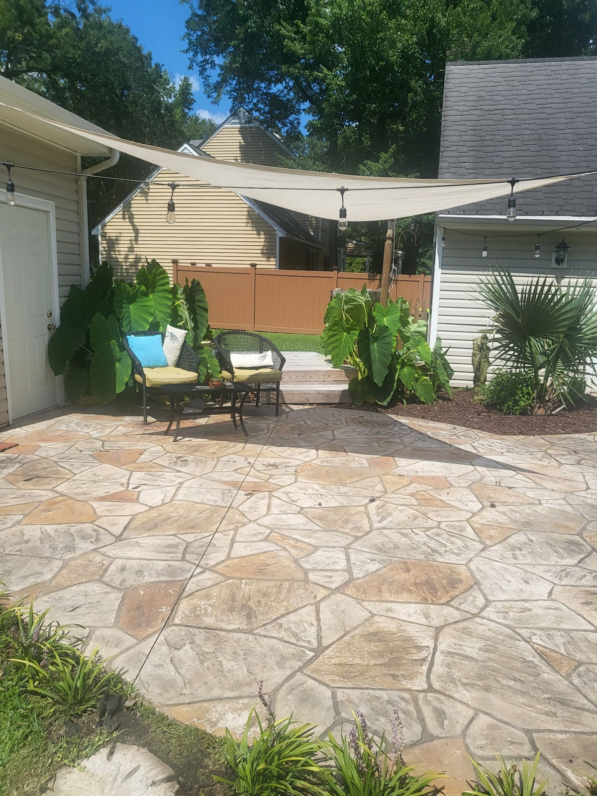 A spacious outdoor patio is highlighted by a stone-patterned surface, adorned with tropical plants. Two comfortable chairs, accompanied by colorful cushions, are positioned beneath a shade sail, creating a relaxing area for outdoor enjoyment. Soft lighting enhances the tranquil setting.