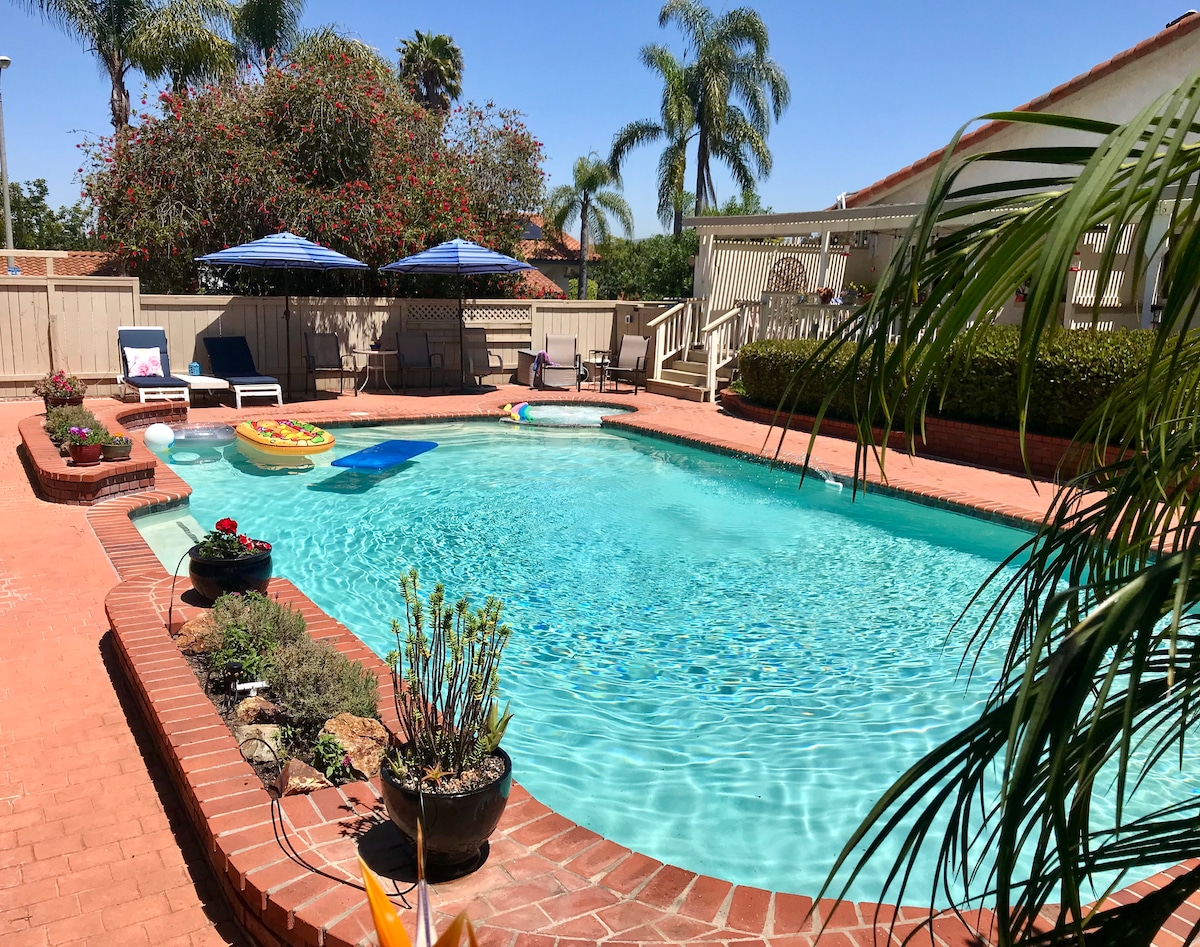 A clear blue pool is surrounded by a brick patio, with lounge chairs and umbrellas providing shaded seating. Lush greenery and potted plants add natural elements to the space, complemented by a decorative garden area visible in the background.