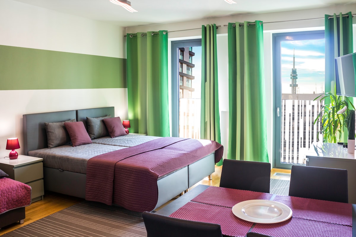 A modern studio apartment features a spacious bed with dark gray bedding and purple accents. Large windows are dressed with green curtains, allowing natural light to illuminate the room. A dining table with black chairs is positioned beside a plant, creating a comfortable living space.