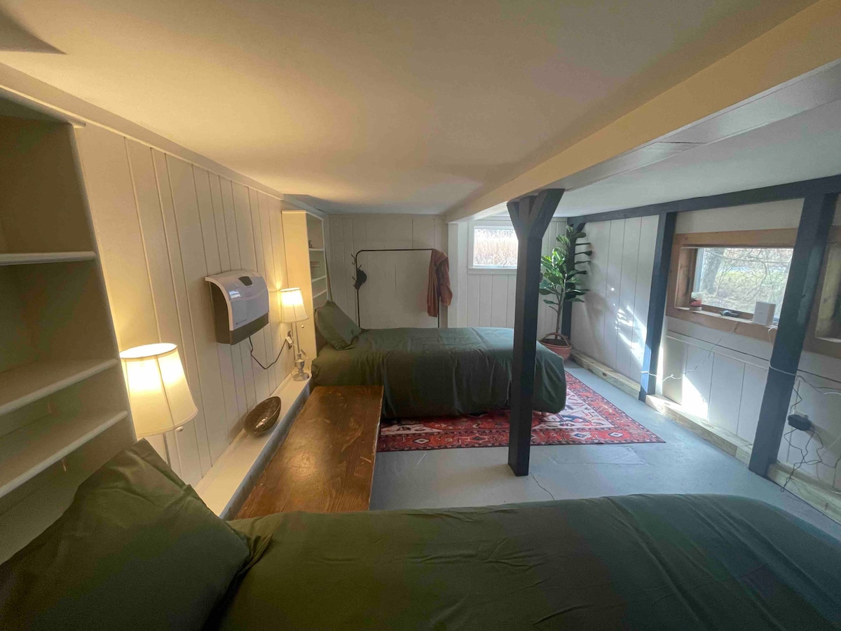 A well-lit sleeping area features two spacious beds covered in dark green bedding. A wooden table is positioned between the beds, with soft lighting provided by lamps on either side. Shelving units and a small indoor plant enhance the tranquil atmosphere.