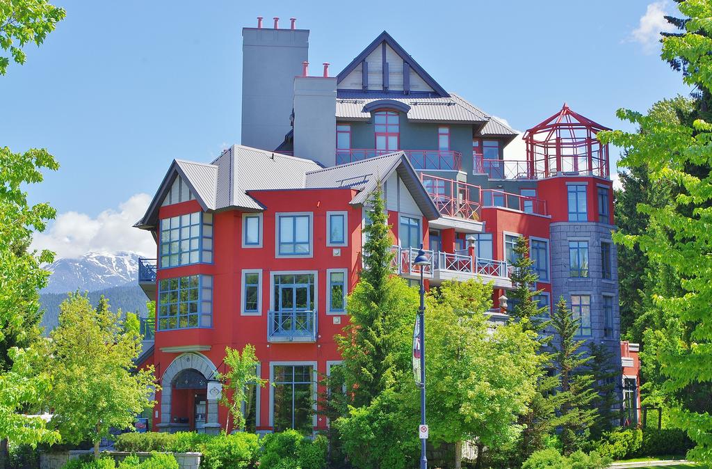 The exterior of a multi-level building is presented, showcasing a blend of colorful red and gray facades. The structure is surrounded by lush greenery and trees, with tall mountains visible in the background under a clear blue sky.