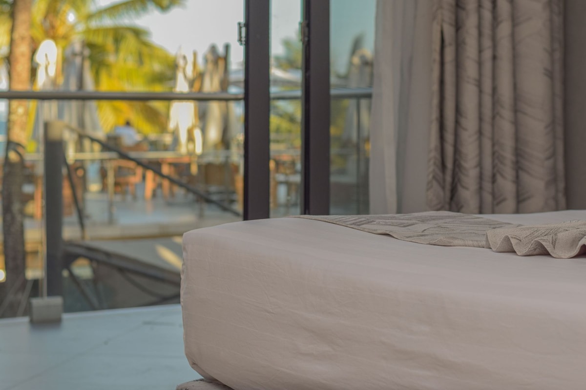 A spacious bed with soft linens is positioned near large glass doors, offering a glimpse of a vibrant outdoor dining area under shaded umbrellas. Lush palm trees are visible in the background, enhancing the tropical ambiance.
