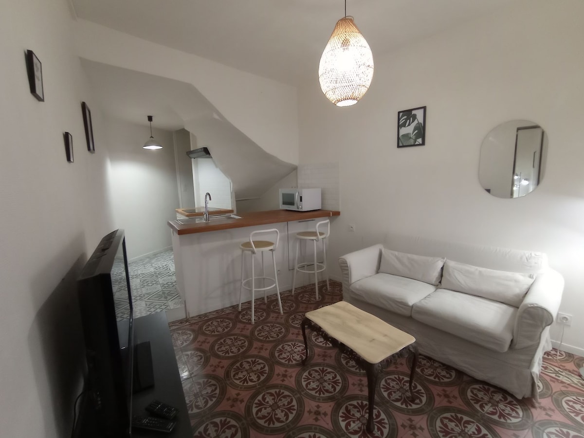 A comfortable living area is visible, featuring a white sofa positioned near a wooden coffee table. A flat-screen TV is present against one wall. The area includes an open kitchen with modern appliances and a circular mirror enhancing the space. Distinctive patterned tiles cover the floor.