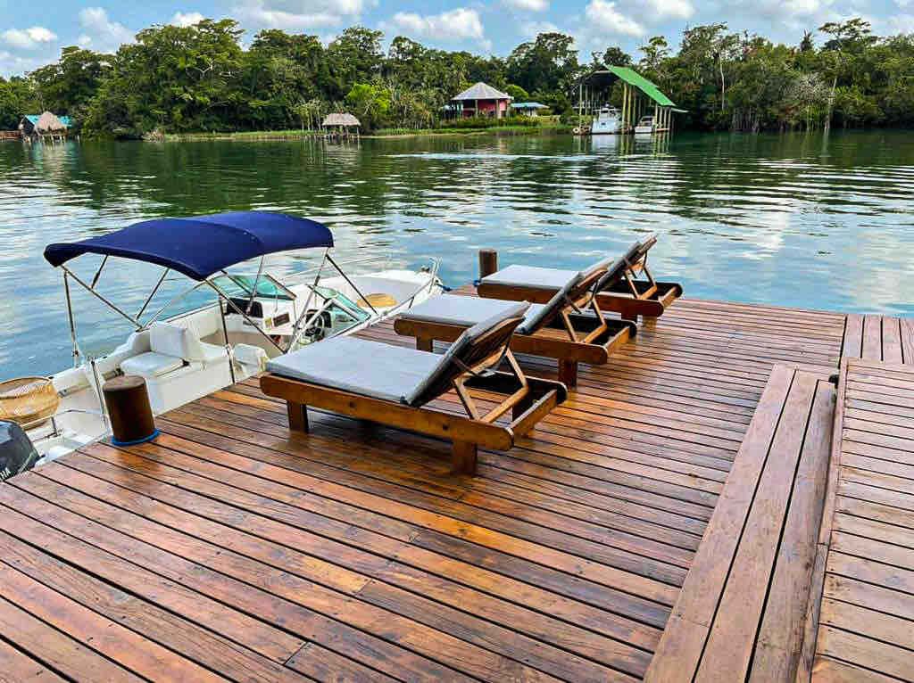 A wooden deck extends over calm waters, furnished with three sun loungers. A canopy provides shade over the loungers, while the surrounding landscape features lush greenery and distant structures, creating a tranquil waterfront setting.