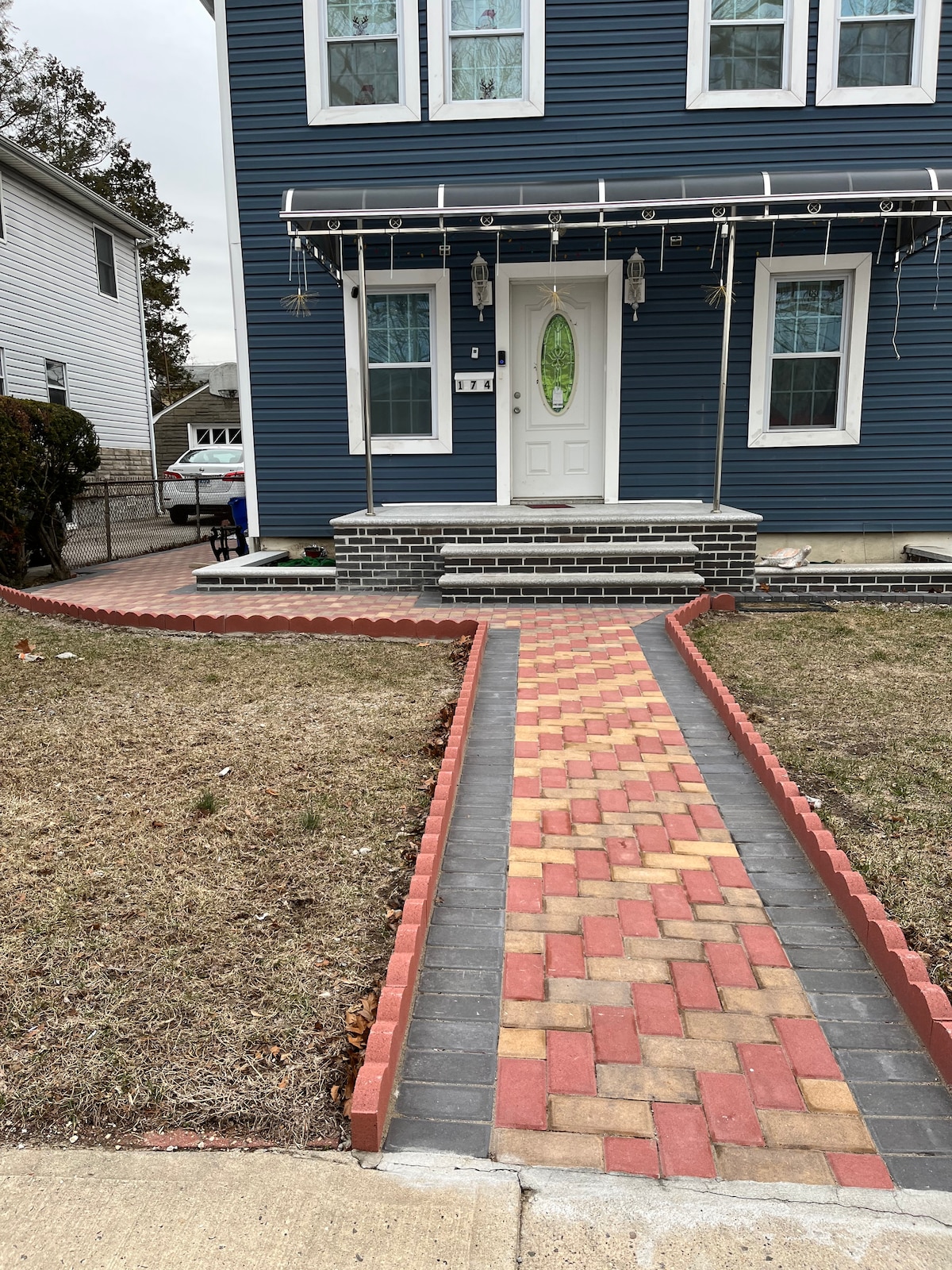 A walkway made of red and gray pavers leads to a blue two-family house. The entrance is framed by two lanterns on either side of a white door, highlighted by a green oval window. Grass borders the path, with homes visible in the background.