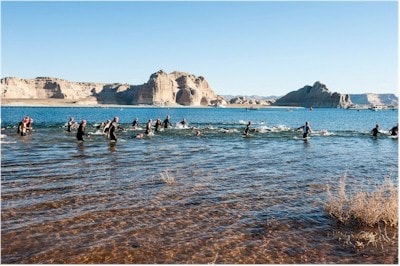 Participants are seen swimming in a calm lake, with a shoreline featuring rocky formations and gentle waves. The backdrop showcases striking cliffs and mountains under clear blue skies, highlighting the natural beauty of the landscape.