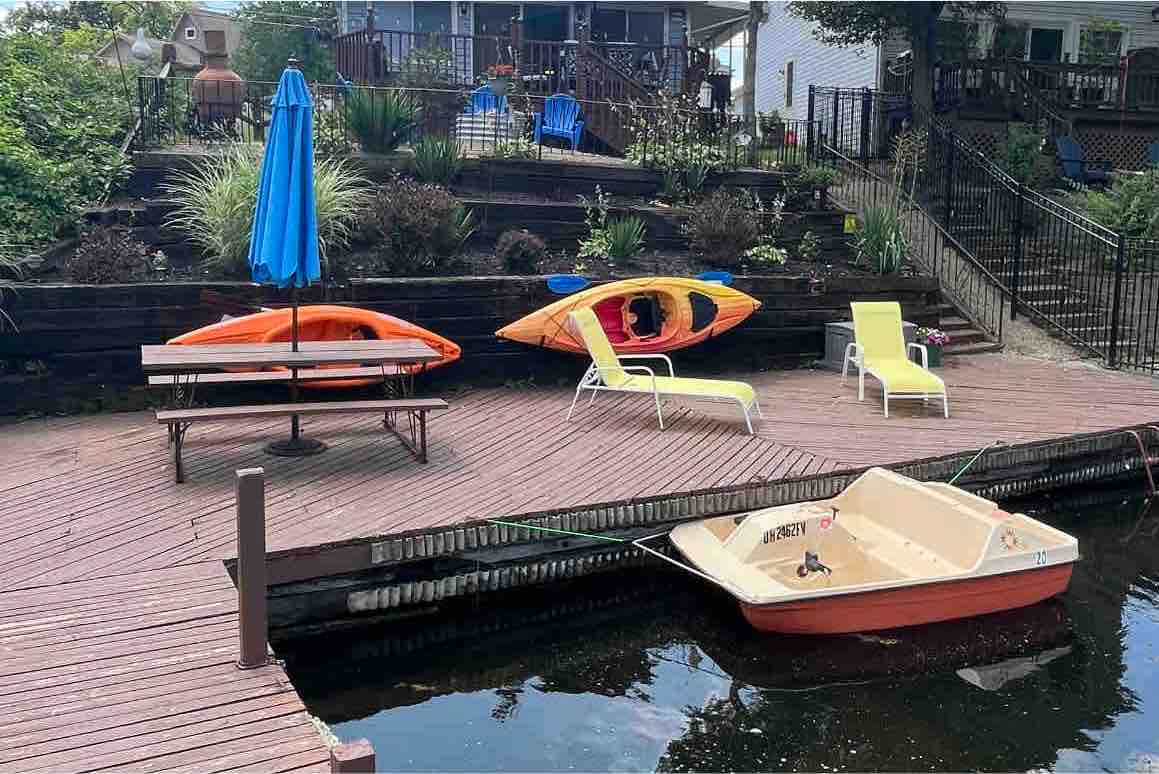 A wooden deck extends to the water, featuring a bright orange kayak and a green kayak positioned alongside a yellow umbrella. Two lounge chairs are arranged for relaxation, with a boat securely tied at the dock, creating a welcoming outdoor space for water activities.