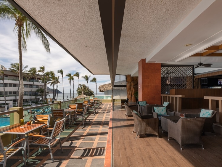 An inviting terrace area with wooden tables and chairs overlooks the pool and beach, surrounded by palm trees. The space features a mix of lounge seating and dining arrangements, providing a relaxed atmosphere with a view of the Pacific Ocean.
