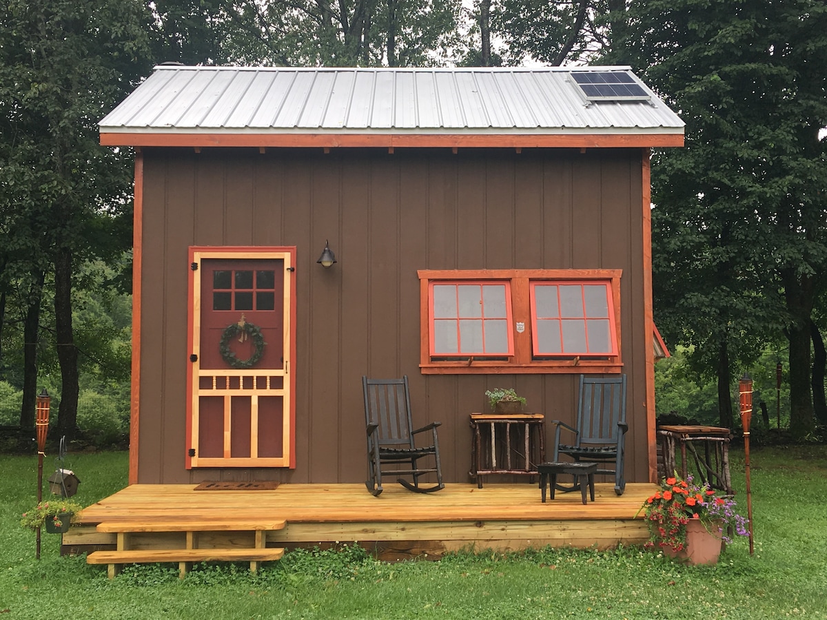 The cabin exterior features a rustic brown facade with bright red trim. A welcoming front door is complemented by two windows, and a simple porch with steps leads to a seating area, furnished with two rocking chairs and a small table. Flower pots add a touch of nature.