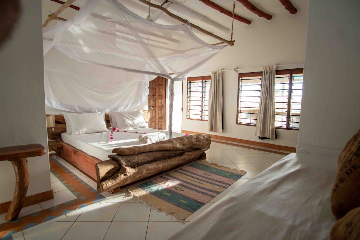 A spacious bedroom features a large bed adorned with white linens and surrounded by a flowing canopy. Natural light streams through several wooden-framed windows. Decorative elements include a woven rug and a rustic wooden bench, emphasizing a relaxed atmosphere.