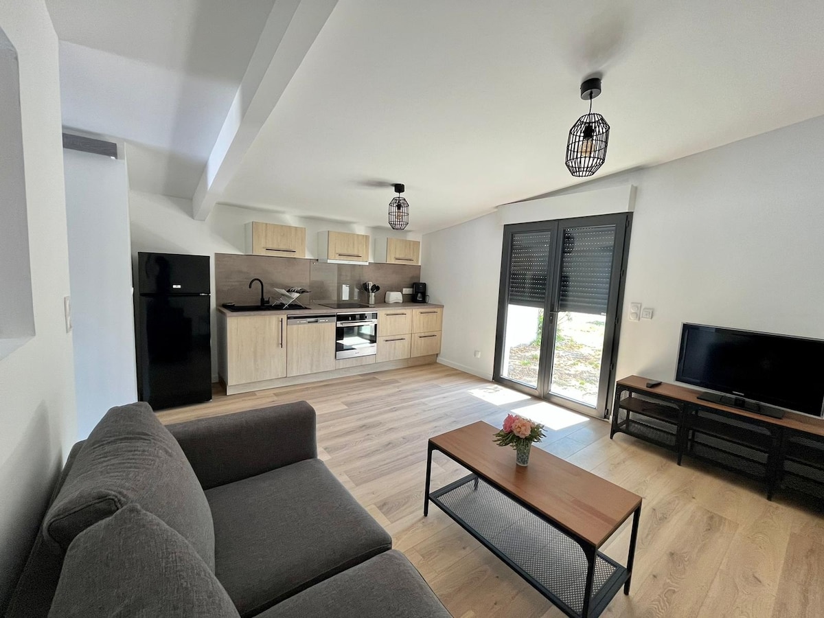 A modern living space features a comfortable gray sofa facing a low wooden coffee table. A flat-screen television is positioned on a media unit. The kitchen area is equipped with light wood cabinetry, a dark refrigerator, and essential appliances, illuminated by pendant lighting.
