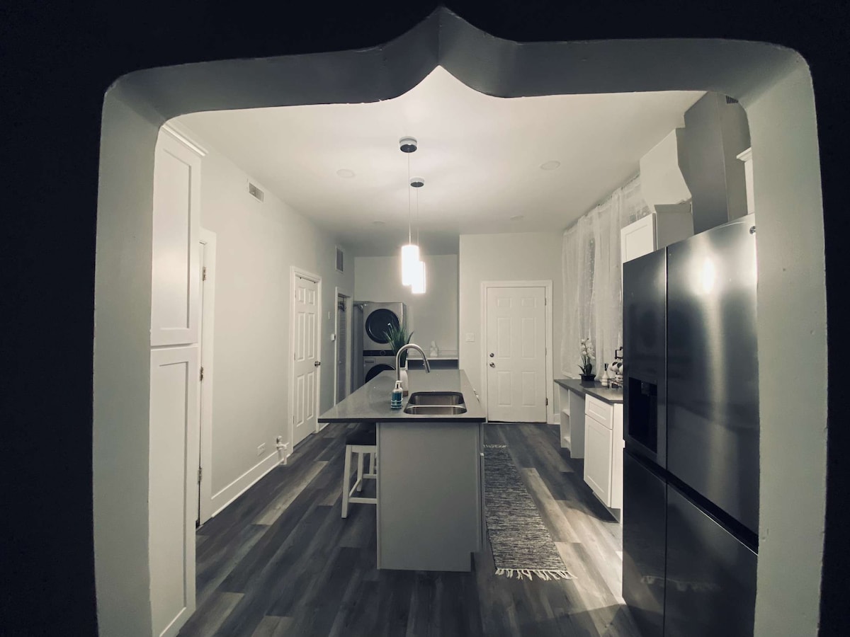 A spacious kitchen is seen through an archway. Bright white cabinets contrast with the dark countertops. An island with a sink is centrally positioned, surrounded by modern appliances. Natural light filters through a nearby window, enhancing the open and airy layout.