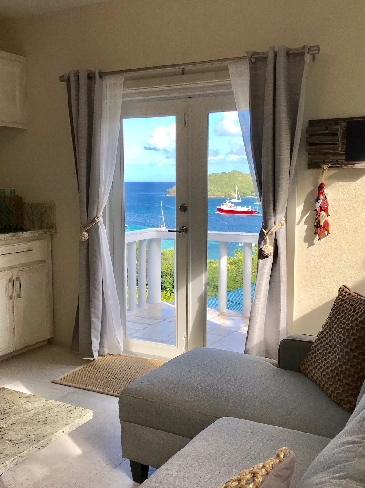 A sliding glass door opens to a balcony with views of Admiralty Bay. Soft gray curtains frame the door, while a light-colored sofa is positioned nearby. The scene includes a glimpse of water and a red and white boat in the distance.