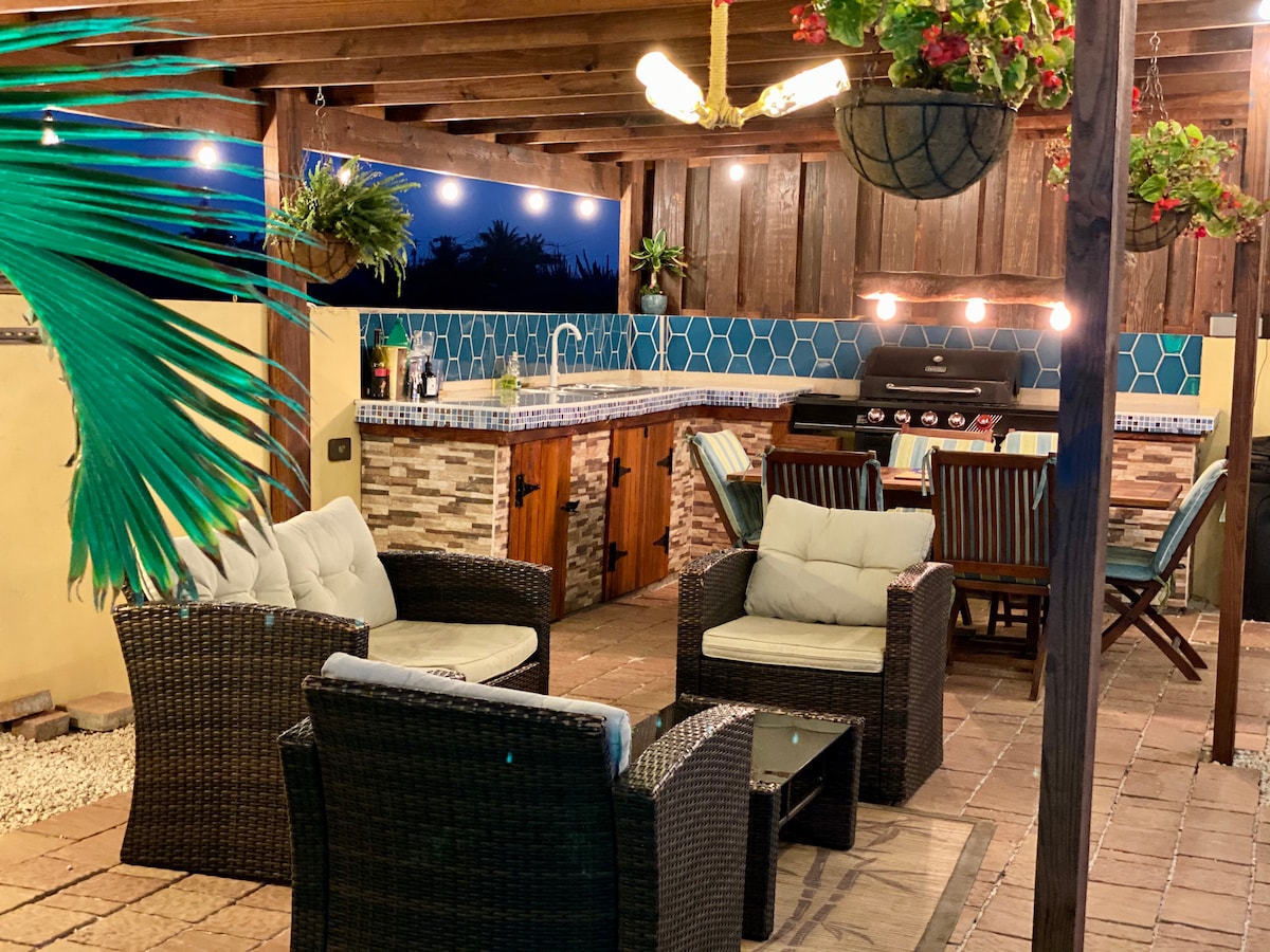 An outdoor patio area is pictured, featuring a comfortable seating arrangement with woven chairs and a table. A grilling station is positioned against the wall, with decorative tile accents. Soft lighting enhances the space, surrounded by tropical plants, offering a relaxed environment for gatherings.