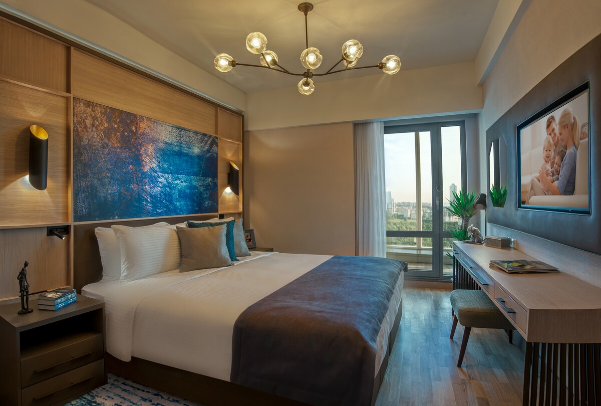A well-appointed bedroom features a spacious bed with soft linens and a decorative throw. A modern chandelier provides ambient lighting, while a large artwork adds a splash of color. A window offers views of the outside, complemented by a sleek desk and decorative elements.