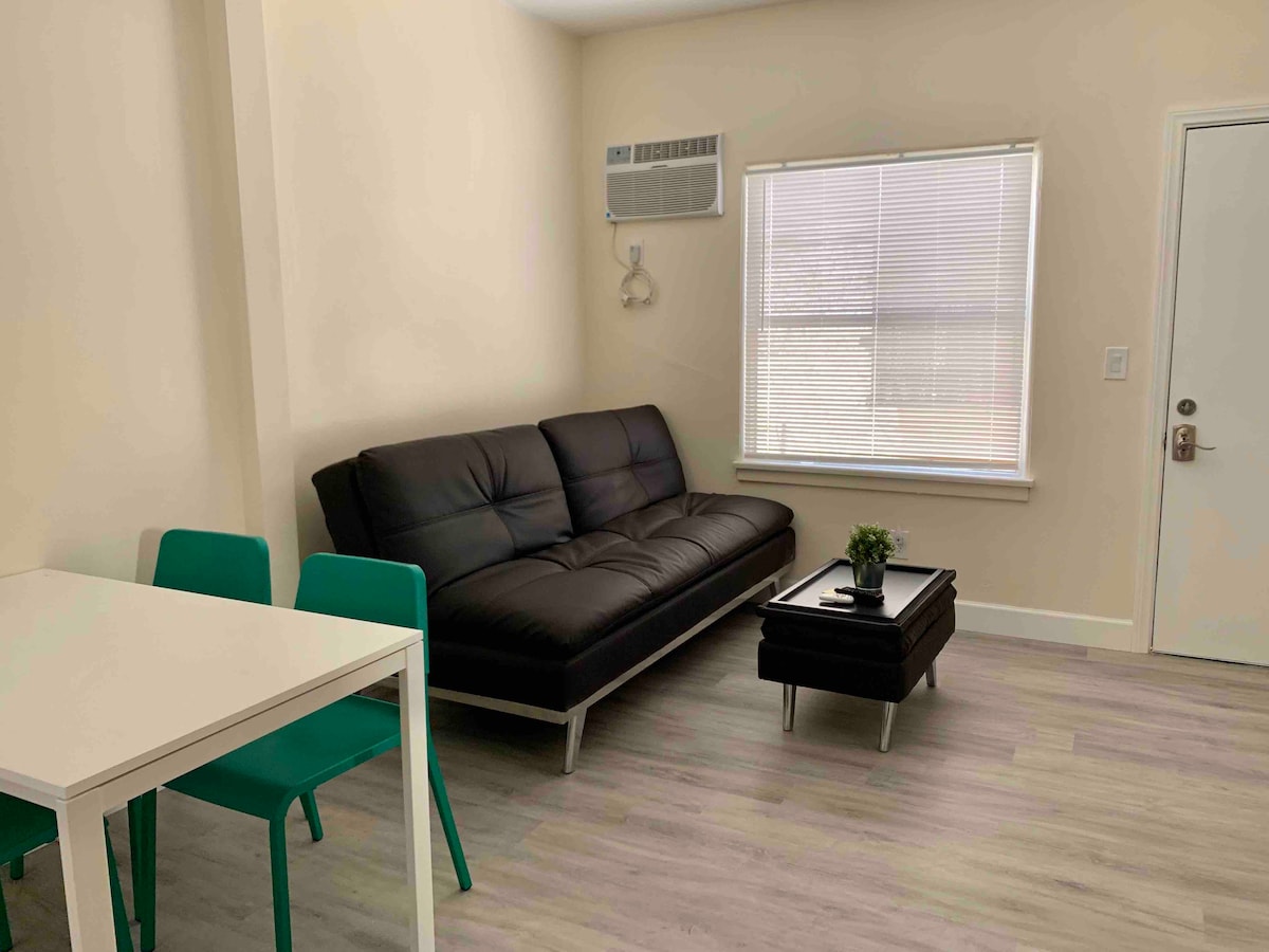 A living area features a sleek black sofa positioned near a window letting in natural light. A small white dining table with green chairs is situated to one side. A coffee table with a small plant is placed in the center, enhancing the space's simplicity.