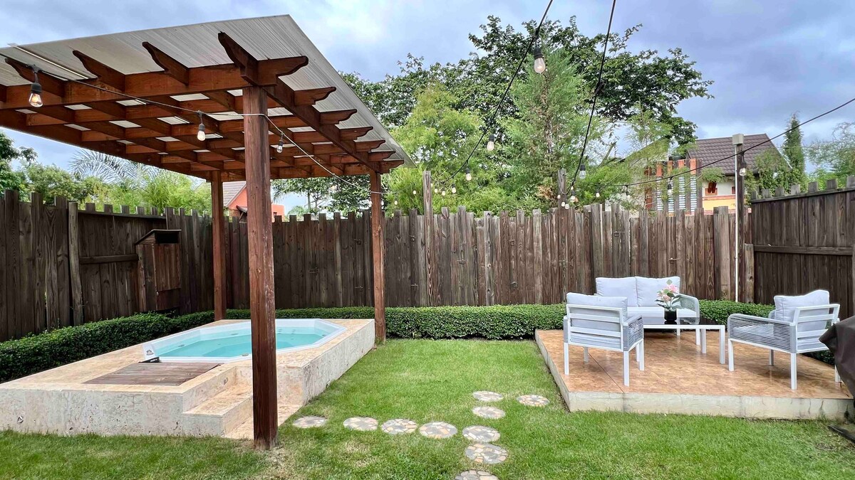 A serene outdoor space is showcased, featuring a heated jacuzzi beside a well-manicured lawn. A wooden pergola provides shade over a seating area, which includes two chairs and a small table. Soft, ambient lighting is arranged overhead, enhancing the peaceful environment.