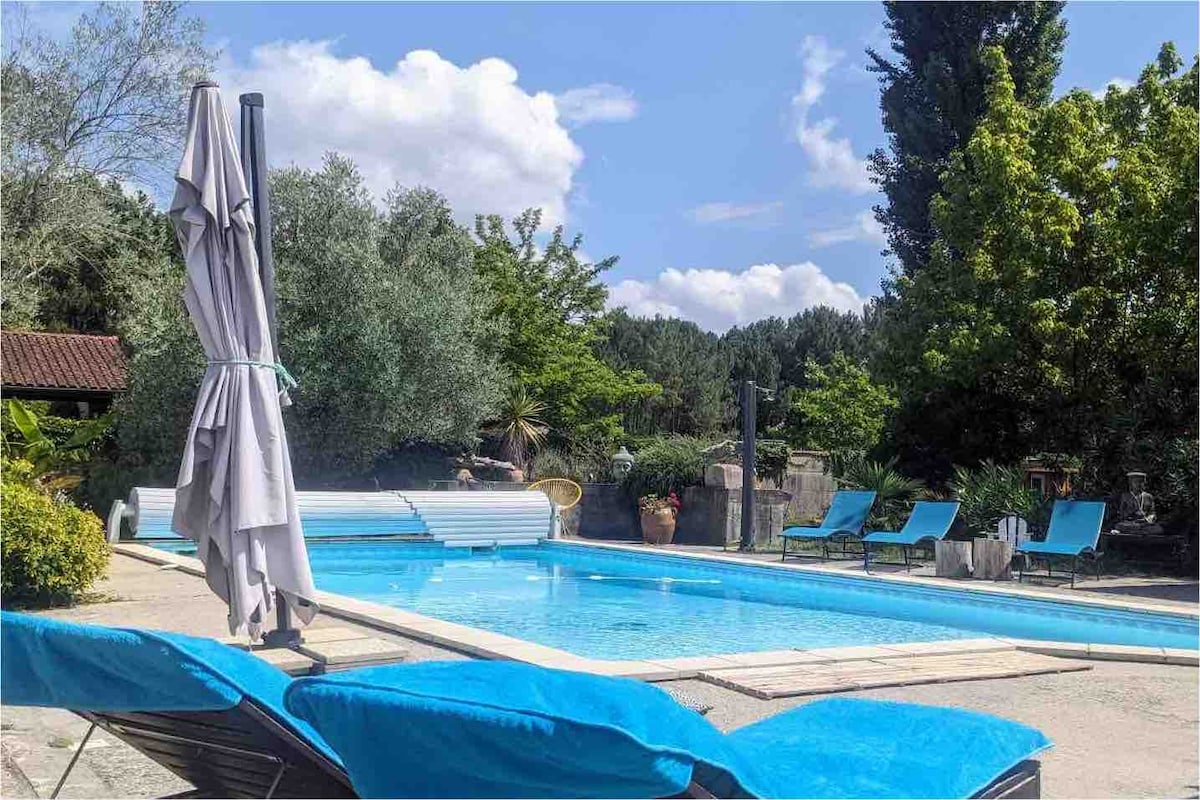 A heated swimming pool measuring 10x5 meters is surrounded by lush greenery. Loungers with bright blue cushions are positioned nearby on a sunlit patio, complemented by a large sun umbrella. The tranquil atmosphere is enhanced by trees and plantings in the background.