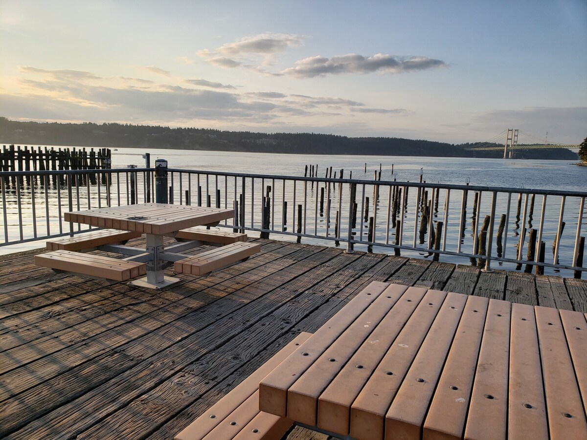Two wooden picnic tables are positioned on a deck overlooking calm waters. The scene features a peaceful reflection of the sky at sunset, with gentle ripples on the surface. In the background, wooden pilings emerge from the water, and a bridge can be seen in the distance.