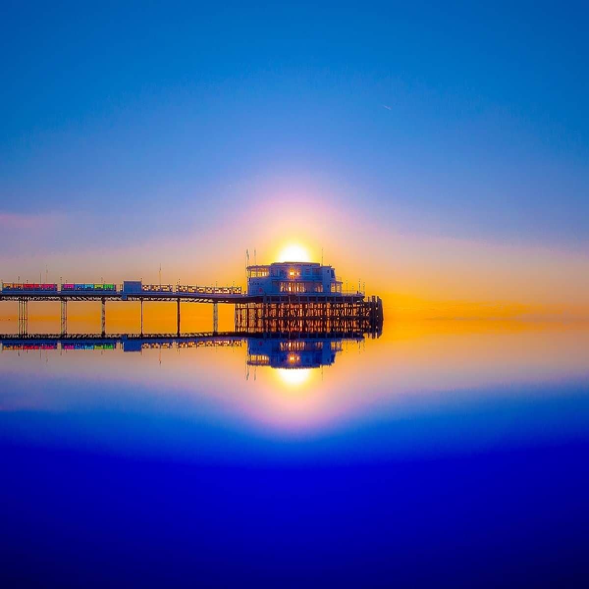 A serene image of a pier extending into calm water, reflecting the warm hues of the sunrise. The soft gradient of blue and orange tones fills the sky, creating a tranquil atmosphere. The structure of the pier is prominently featured against the horizon.
