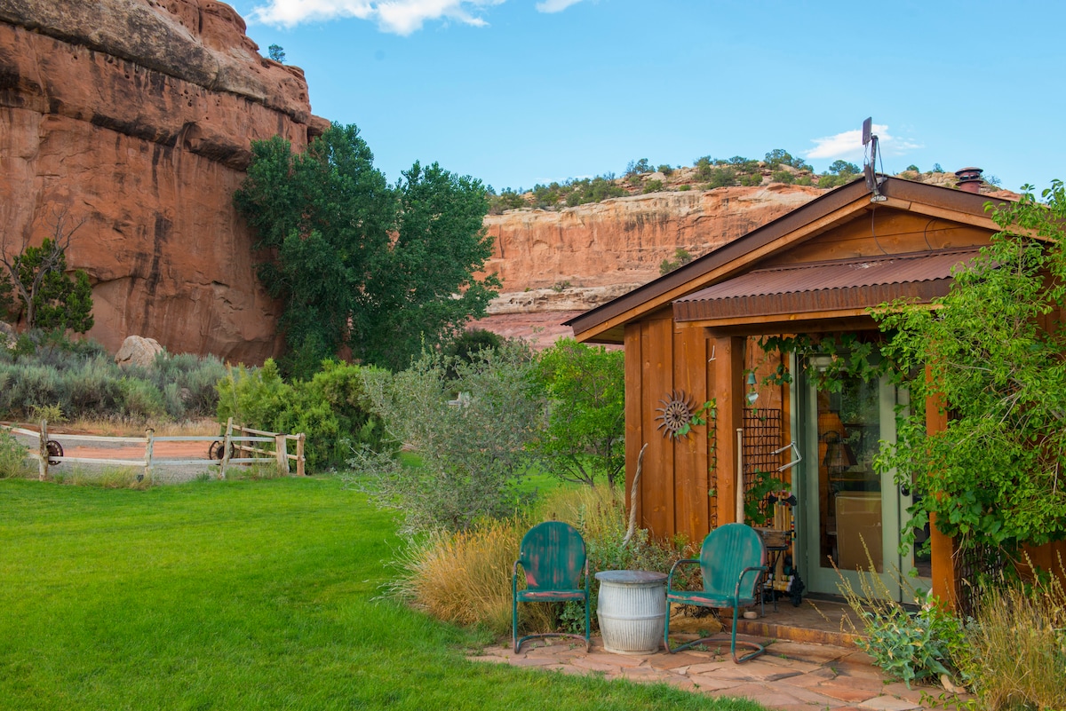 The exterior of a wooden cabin is shown, nestled in a green garden. Two chairs sit in front of the cabin, with a small round table nearby. The backdrop features striking rock formations and lush trees, contributing to a serene setting.