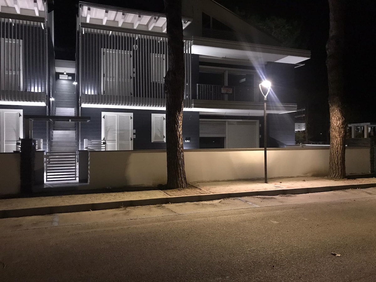 A contemporary two-story building is captured at night, illuminated by exterior lighting. The structure features horizontal wooden slats and multiple balconies, with a paved path leading to the entrance. Tall trees flank the property, creating a serene atmosphere.
