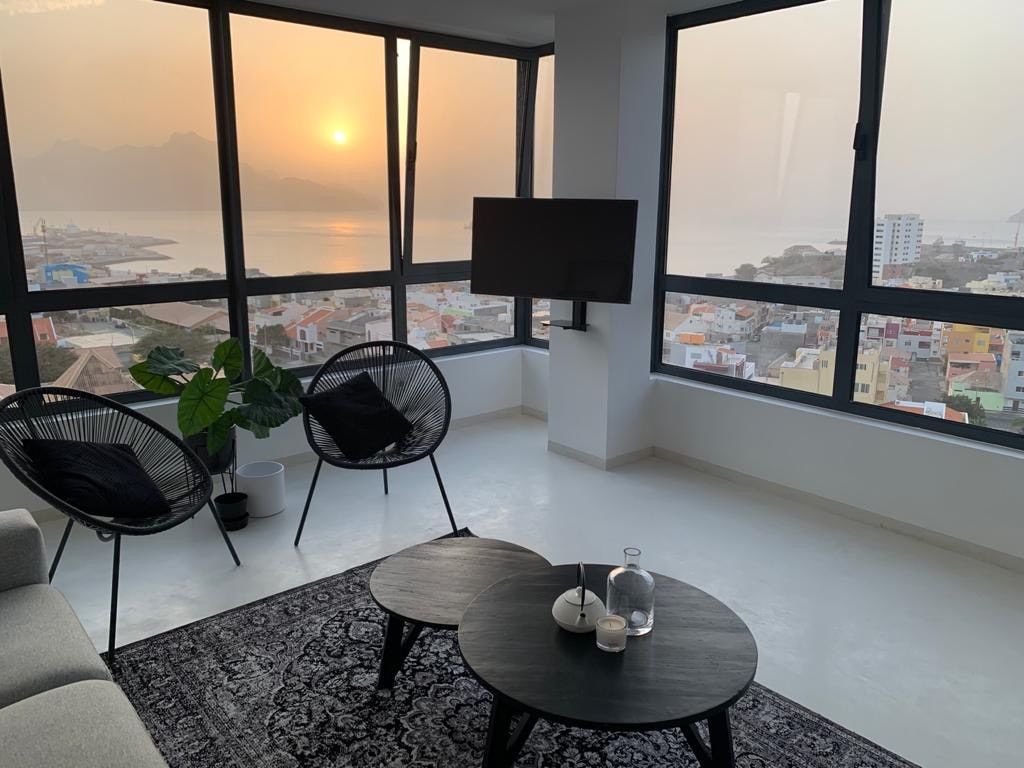 A bright living area features large windows that provide a stunning view of the sunset over the sea and cityscape. Two modern black chairs are positioned beside a round coffee table, complemented by a decorative plant. A flat-screen television hangs on the wall, enhancing the space.