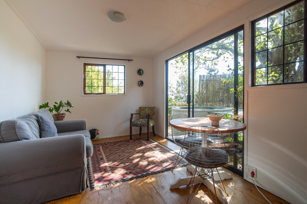 A cozy lounge area features a comfortable grey sofa and a dining table, bathed in natural light from large windows. A colorful chair is tucked next to a potted plant, with a patterned rug adorning the wooden floor. Views of greenery can be seen outside.