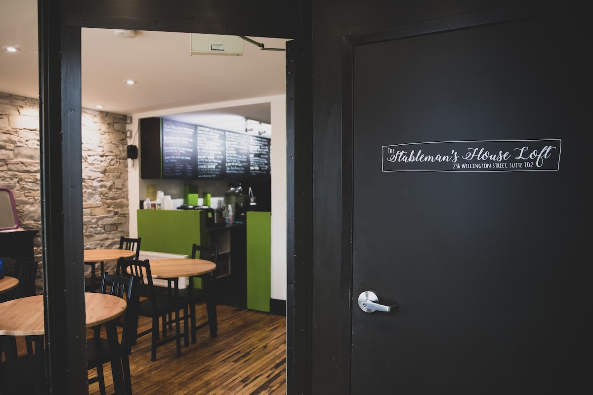A welcoming café area is visible through a doorway. Simple wooden tables and black chairs are arranged against stone walls. Green accents complement a menu board displayed in the background, creating a bright and casual dining atmosphere. The entrance features a sign identifying The Stableman's House Loft.