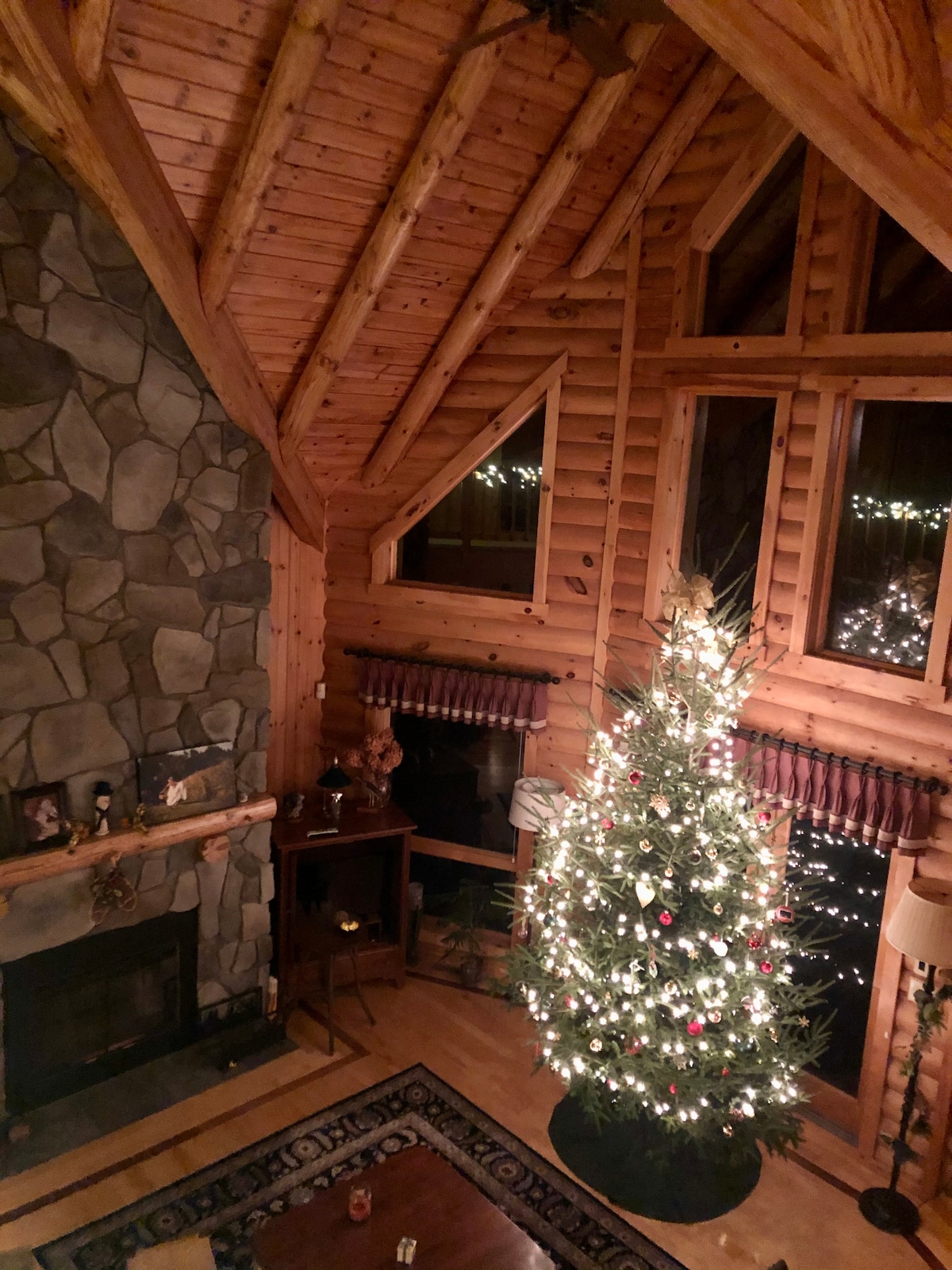 The great room features a high, wooden ceiling with exposed beams. A full-height stone fireplace is framed by a beautifully decorated Christmas tree, adorned with lights and ornaments. Large windows allow soft light to fill the space, enhancing the rustic charm of the log home.