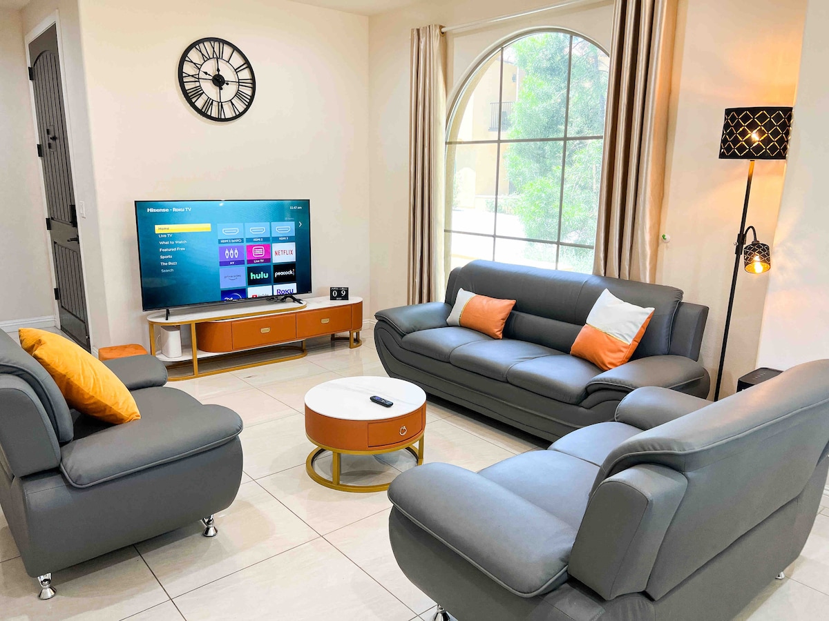 The living room features a modern design with two gray leather sofas, one adorned with orange accent pillows. A flat-screen TV is mounted on the wall, and a decorative clock adds character. The large window allows natural light to fill the space, complemented by soft curtains.