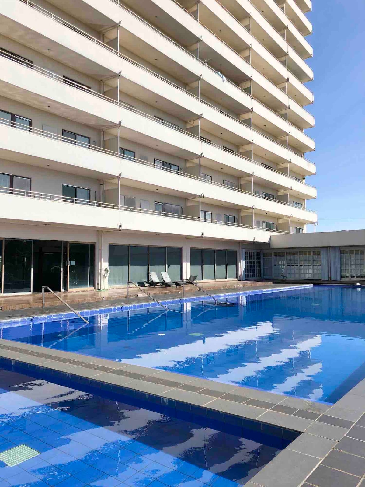A modern high-rise building is featured, with multiple balconies overlooking a clear blue swimming pool. The pool area includes steps leading into the water, with lounge chairs positioned along the edge, inviting relaxation under the open sky.