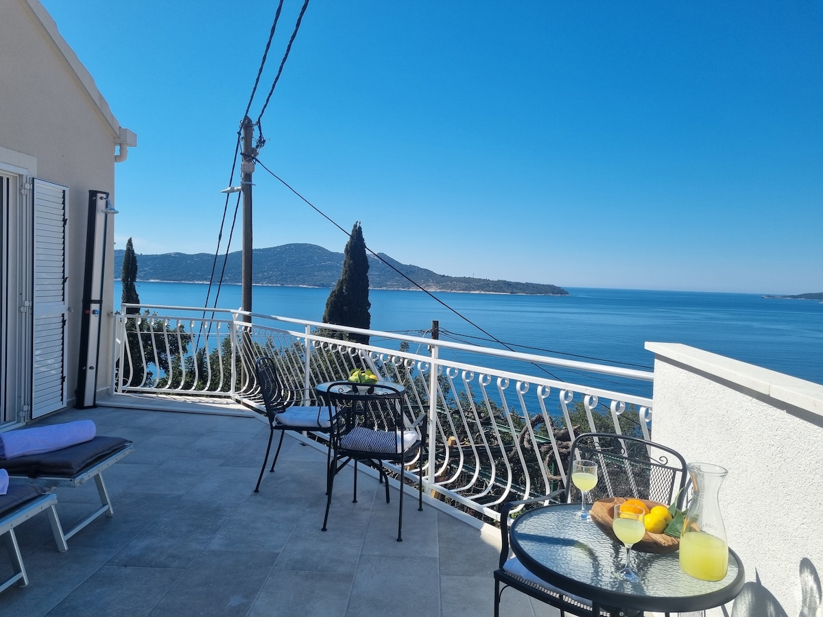 A terrace is displayed with a stunning sea view, featuring two metal chairs and a small round table set with refreshments. Sunbeds are positioned nearby, providing a serene spot for relaxation under clear blue skies.