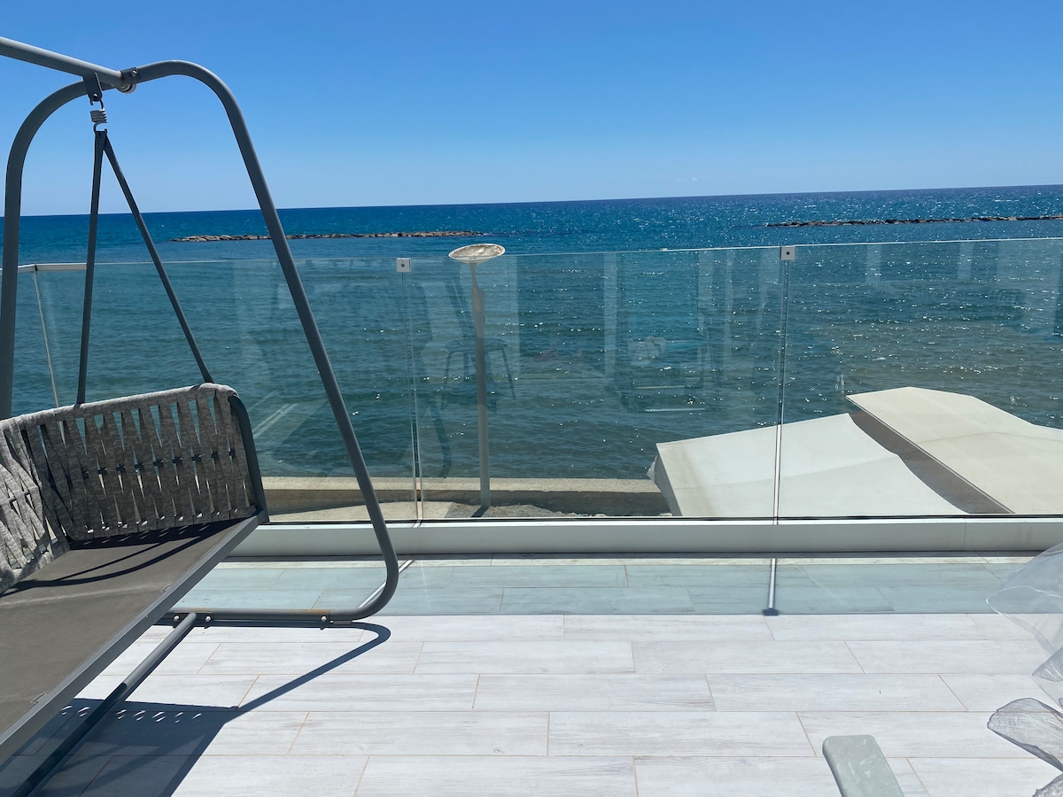 A wooden swing chair is positioned on a balcony, facing the tranquil sea. The view includes expansive water and a distant shoreline, separated by a glass barrier that offers unobstructed sightlines. The surface below is paved with light-colored tiles, reflecting sunlight.