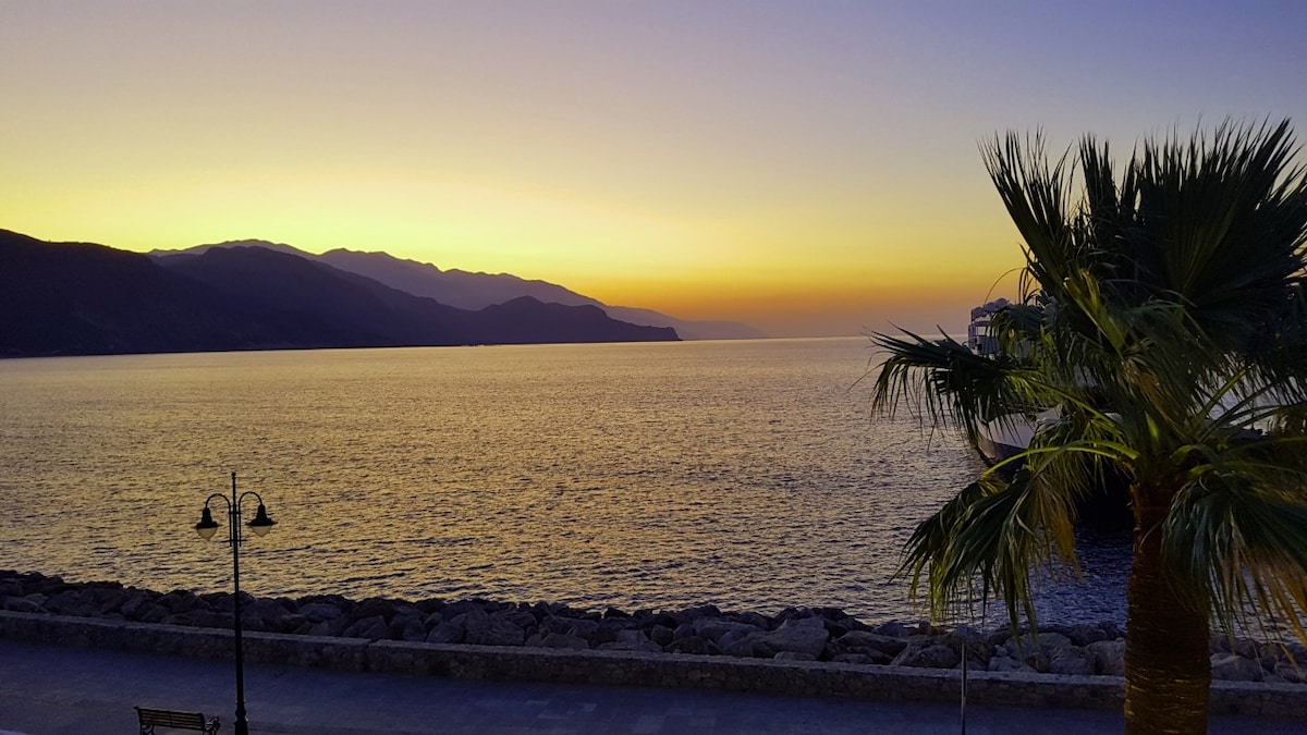 A serene seascape captures a vibrant sunset over the water, with hues of orange and yellow blending into a twilight sky. Silhouetted mountains outline the horizon, while a palm tree and a street lamp frame the scene, offering hints of the coastal landscape.