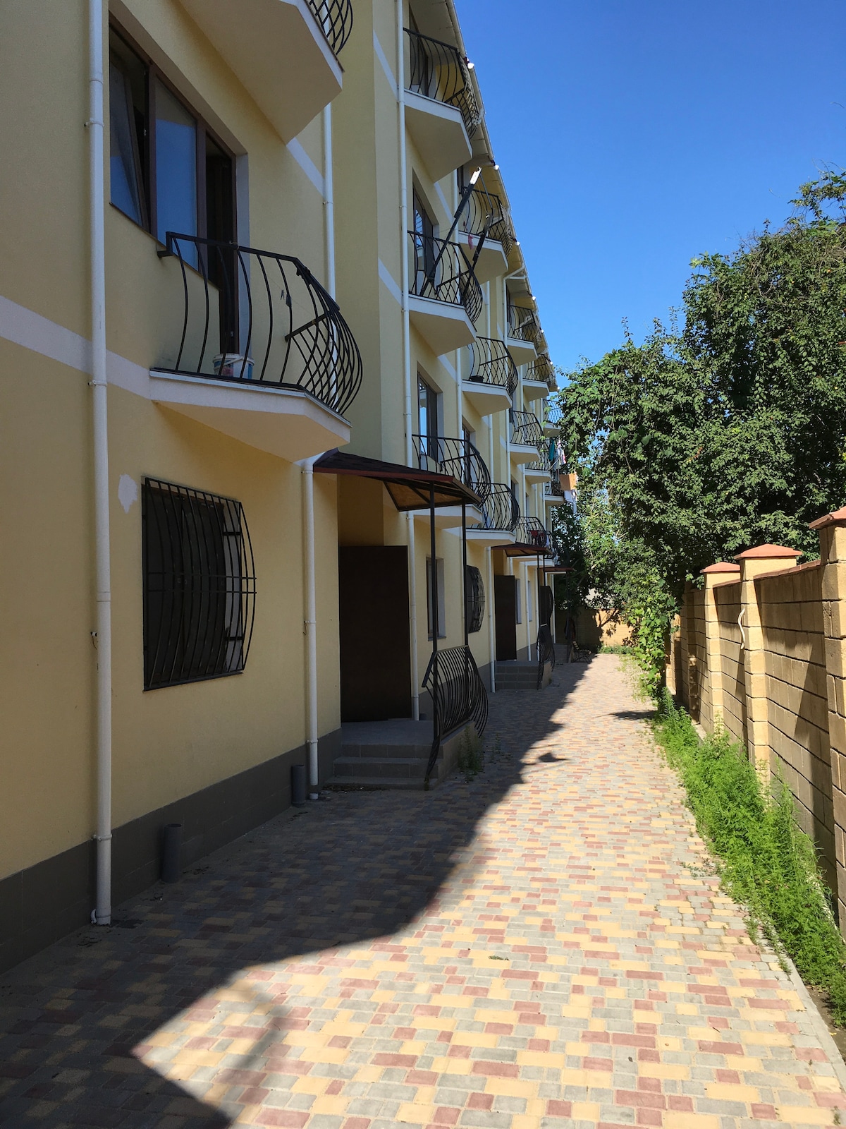 A narrow, paved pathway is shown next to a yellow building with several balconies. The path is lined with green shrubs and trees, providing a natural contrast to the structure. Sunlight brightens the scene under a clear blue sky.
