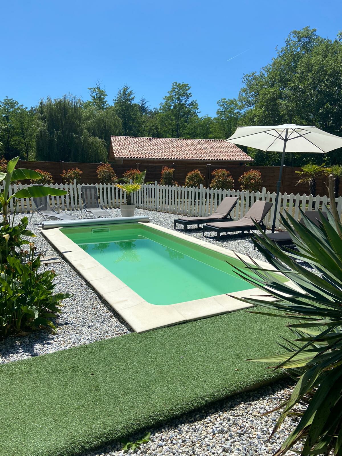 A private pool is surrounded by a well-maintained garden, featuring lounge chairs and an umbrella for shade. The pool is framed by lush greenery and decorative stones, creating a serene outdoor space for relaxation.