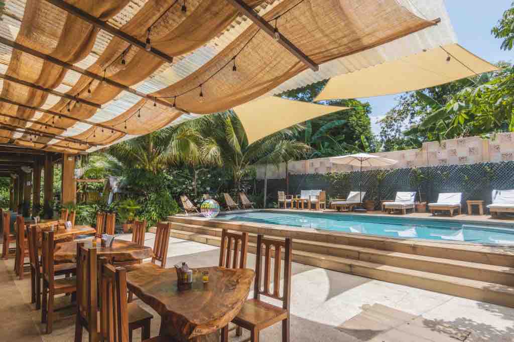 An outdoor dining area features wooden tables surrounded by chairs beneath a canopy of woven materials and shade sails. A clear blue pool is positioned nearby, with lounge chairs arranged around it, set against a backdrop of lush greenery and palm trees.