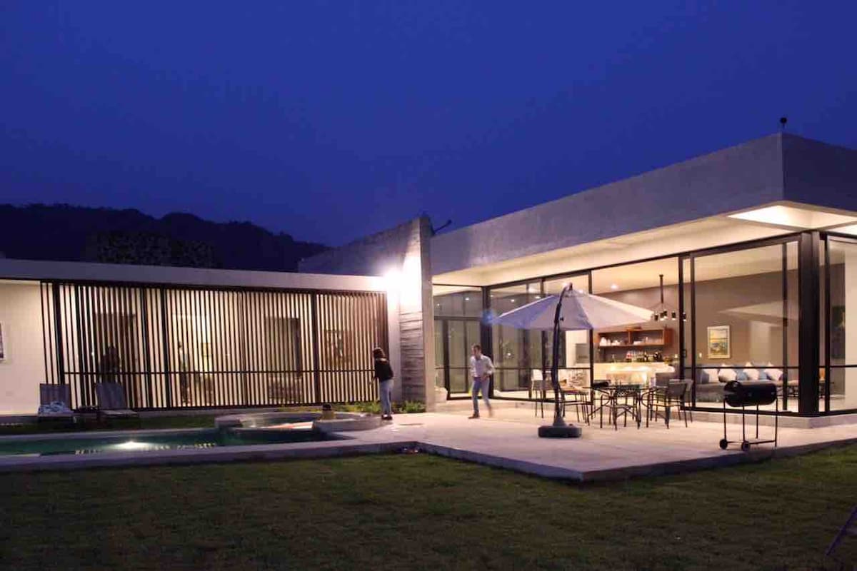 The modern single-story house is illuminated against a twilight sky, showcasing large glass walls that open to a patio. A shaded umbrella provides seating area protection, and a pool is visible in the foreground, surrounded by a neatly maintained lawn.