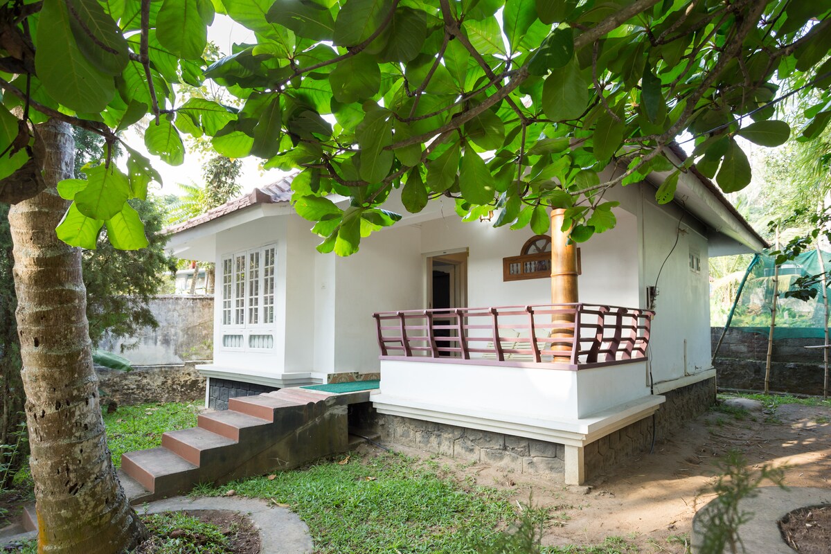 A charming exterior view of a villa is captured, showcasing a porch with wooden railings. Steps lead up to the entrance, surrounded by lush greenery and a well-maintained lawn. Sunlight filters through the leaves of nearby trees, creating a serene outdoor setting.