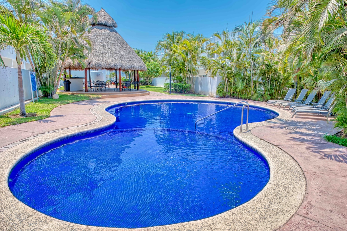 A shared pool is surrounded by lush greenery and palm trees, featuring a thatched-roof palapa nearby. Lounge chairs are arranged along the poolside, inviting relaxation. The pool's unique shape is complemented by a clear blue water surface reflecting the sunlight.