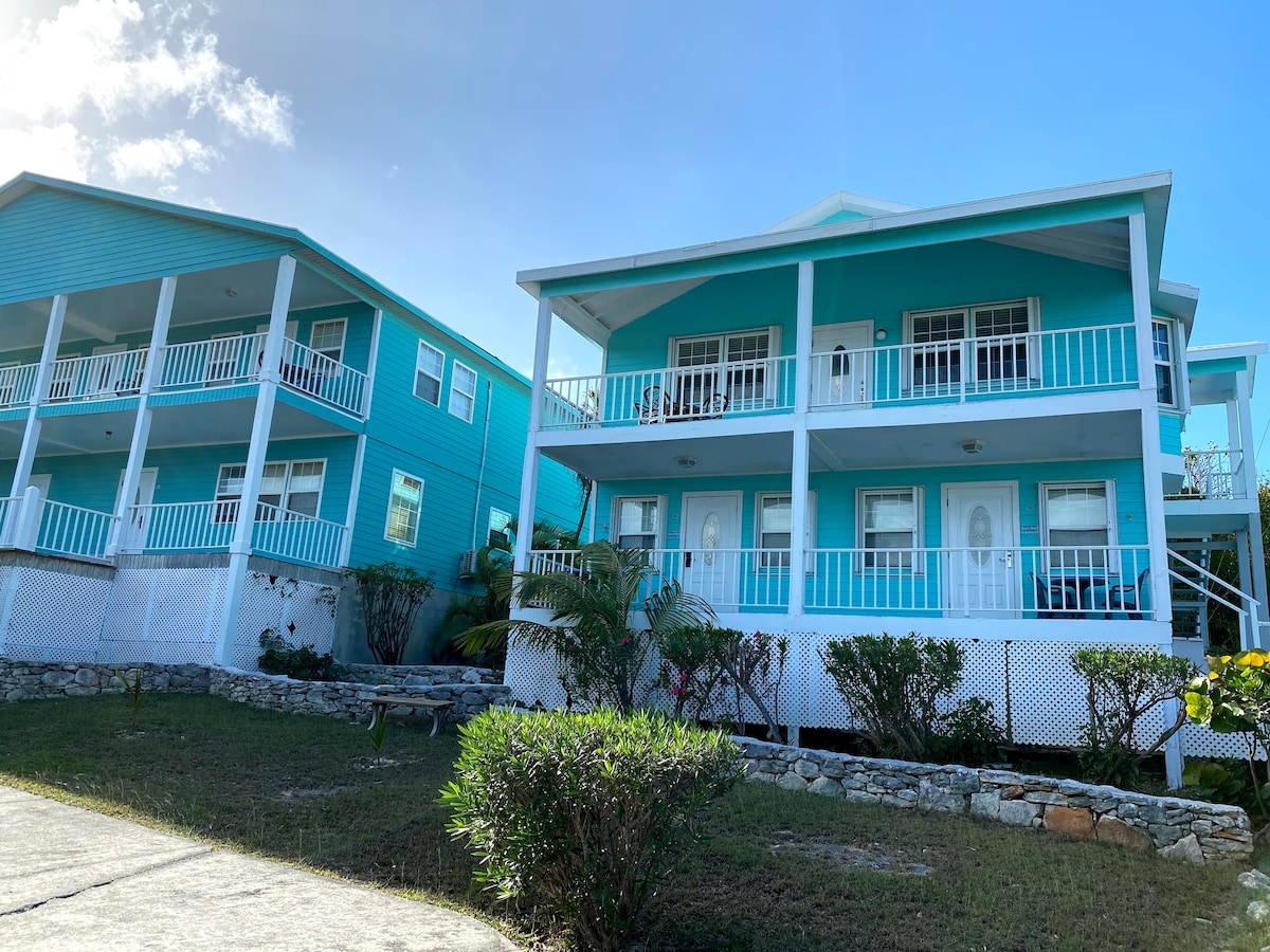A vibrant turquoise building is displayed under clear blue skies. The two-story structure features white railings and expansive balconies. Lush greenery surrounds the property, and a stone border frames the landscaped areas, creating a welcoming appearance.