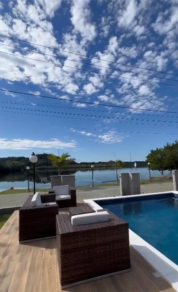An inviting outdoor space features a swimming pool bordered by a wooden deck. Two lounge chairs with white cushions are positioned nearby, offering views of the calm canal and the sky above. Lush greenery is visible beyond the water.