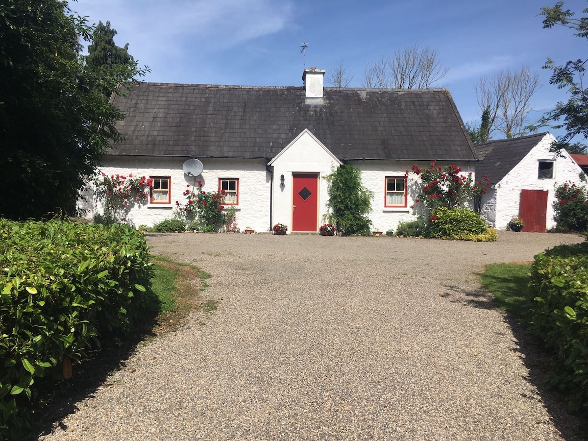 A charming cottage with a white exterior and red accents is situated along a gravel pathway. Lush greenery surrounds the home, enhancing its appeal. A small outbuilding is visible on the right, contributing to the rustic setting amid a clear blue sky.