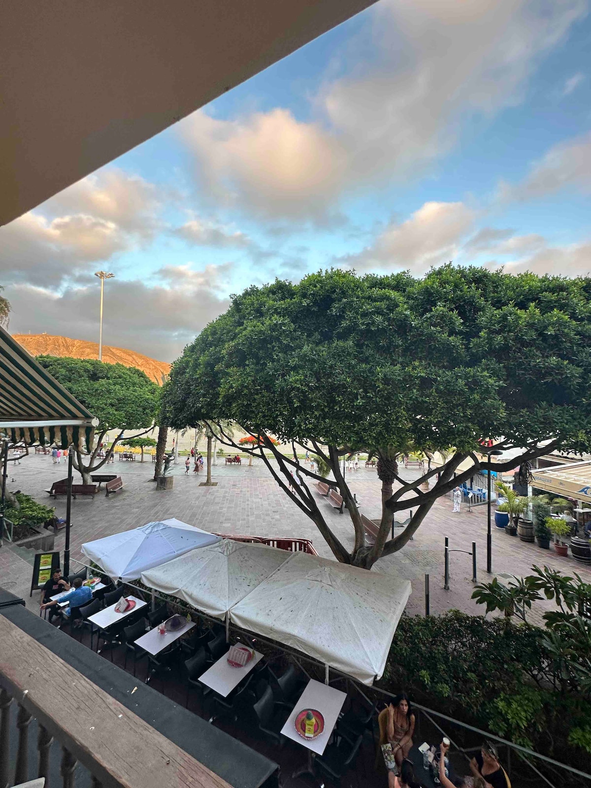 A view from the balcony showcases a vibrant outdoor area beneath a large tree. Multiple tents provide shade over tables where guests are seated. The sky features soft clouds, hinting at a pleasant atmosphere in the central location.