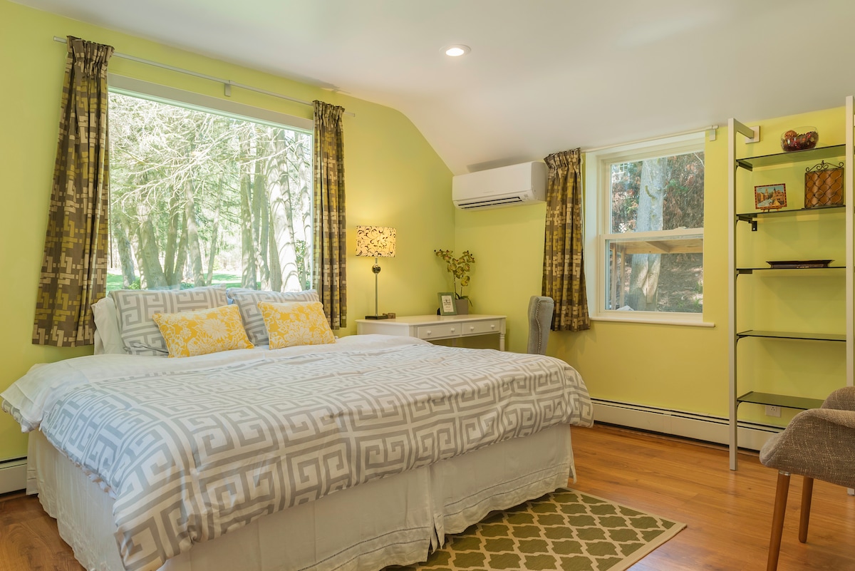 A bright bedroom features two windows overlooking a private outdoor area, allowing natural light to fill the space. A spacious bed is adorned with patterned bedding and yellow accent pillows. A small desk and chair are positioned near the window, complemented by a modern shelving unit.
